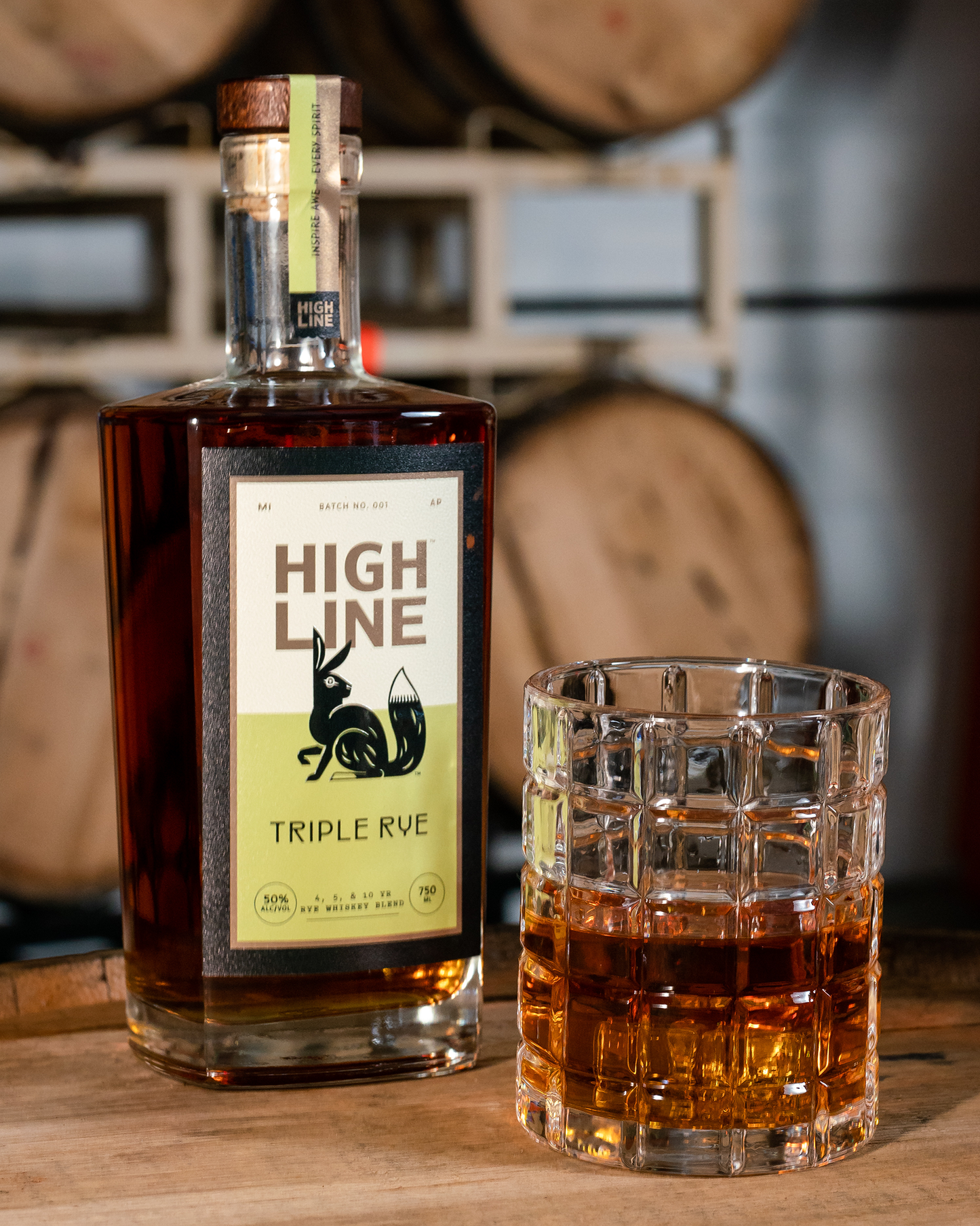 High Line Triple Rye whiskey bottle and glass on a wooden surface with barrels in the background